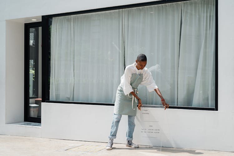 Young Black Female Barista In Uniform Standing Near Signboard Against Cafe