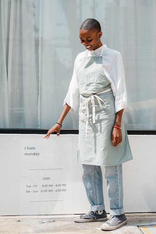 Full length of optimistic young bald black woman in uniform standing near signboard with i hate monday and open inscriptions while readying cafe for opening in morning