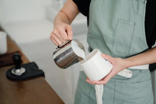 Free From above of crop young anonymous person in apron pouring milk into white mug while preparing aromatic latte Stock Photo
