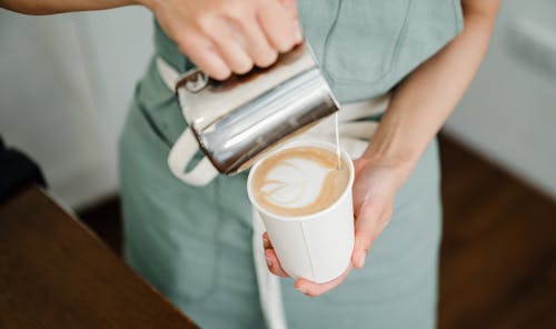 High angle of crop anonymous young barista in apron pouring milk into disposable cup and making latte art