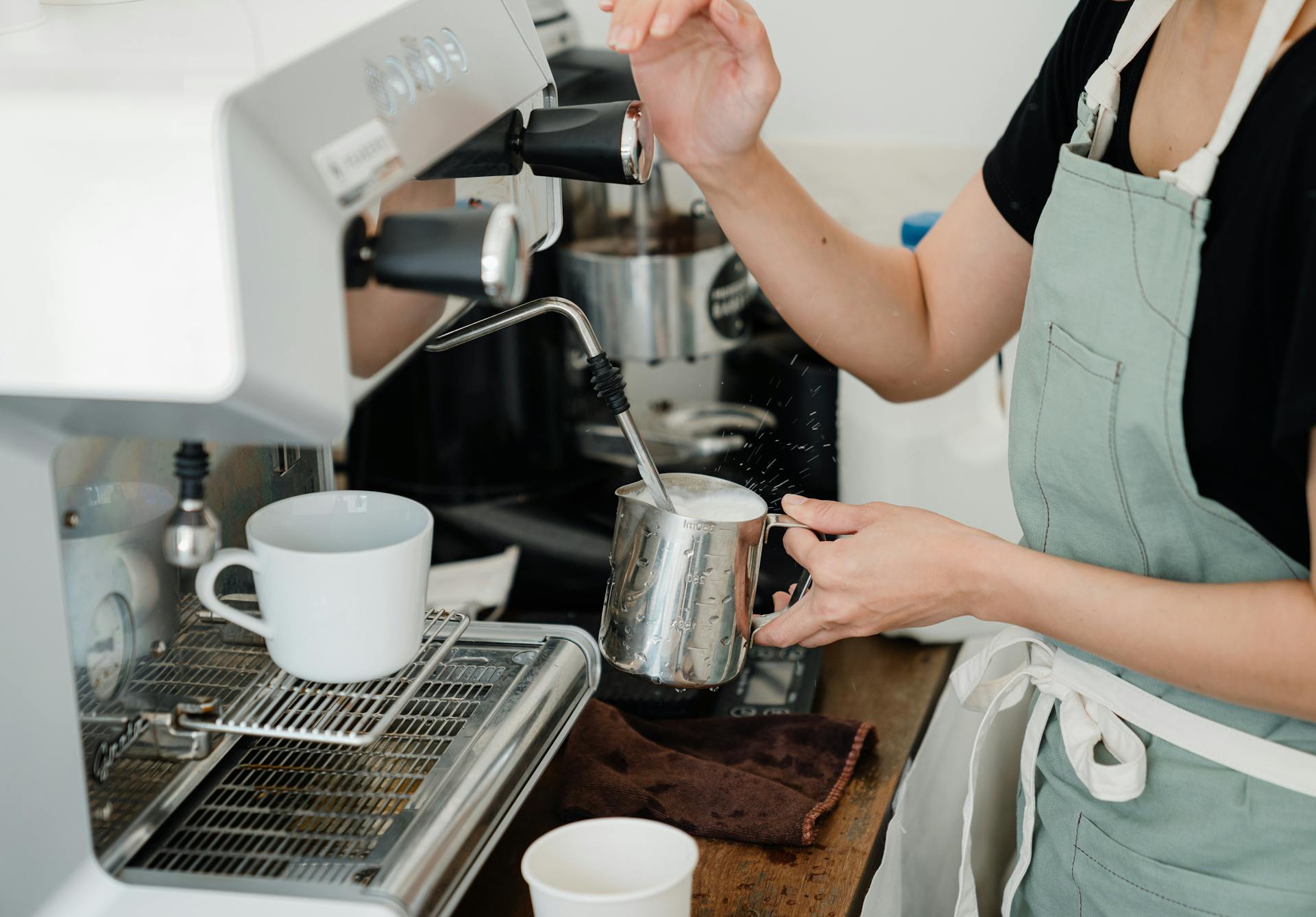 Crop faceless young female barista in apron steaming milk in steel pot using modern cappuccino machine
