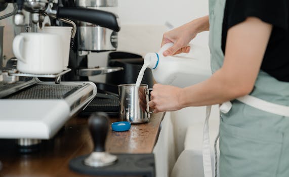 Close-up of a barista pouring milk into a frothing pitcher beside a coffee machine in a café setting.