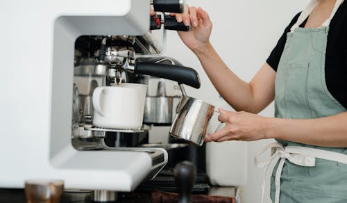 Crop faceless young female barista in apron steaming milk at modern coffee machine while preparing delicious latte