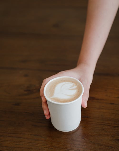 From above of anonymous young barista hand serving delicious cappuccino with milk foam on wooden table