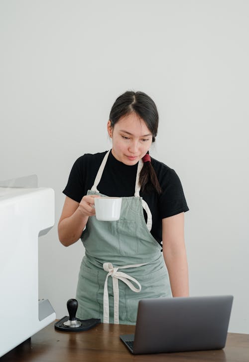 Young Asian female coffee shop worker with coffee in hand watching TV series on laptop while standing at wooden table during break