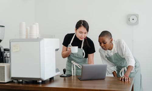 Multiethnic female baristas watching video on laptop with surprised faces while enjoying free time during coffee break at work near table with coffeemaker