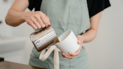 Free Crop unrecognizable barista in apron pouring milk from steel pitcher to white ceramic cup while preparing aromatic coffee in modern cafe Stock Photo