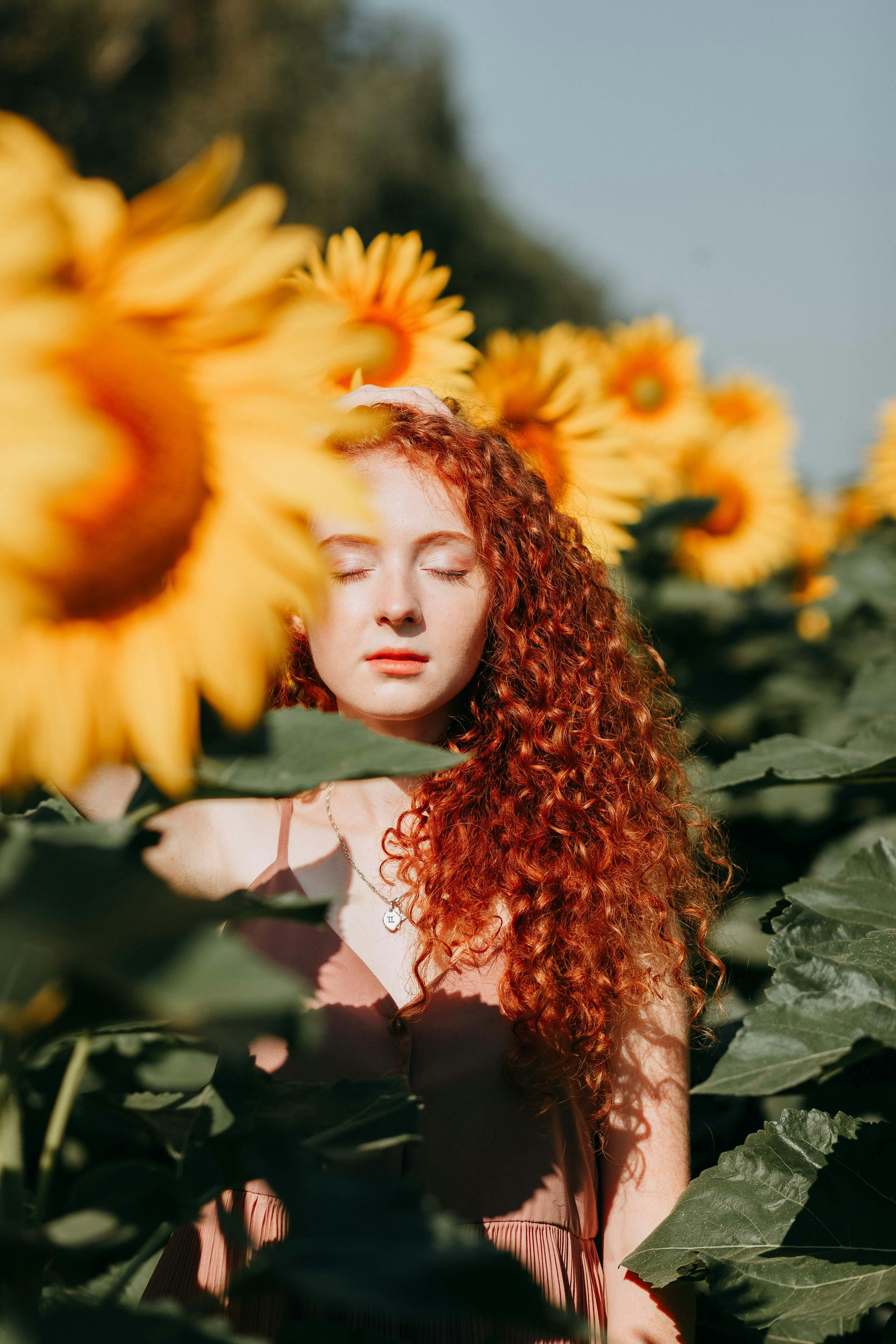 Serene woman with curly hair stands among sunflowers, eyes closed, embracing the warmth of summer sunlight.
