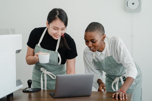 Positive young Asian and African American female baristas in aprons smiling and looking at laptop screen while standing near table and enjoying break at work