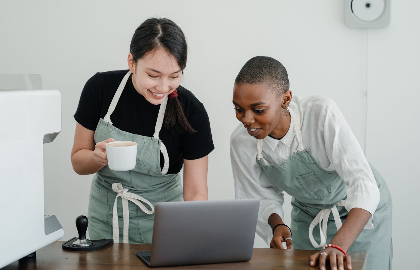 Two baristas smiling while browsing on a laptop in a coffee shop.
