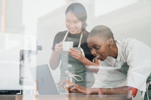 Through glass of happy young diverse female baristas using laptop to type message and smiling while standing at table during break