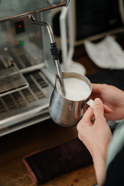 Crop cafe worker using coffeemaker to whip milk in pitcher