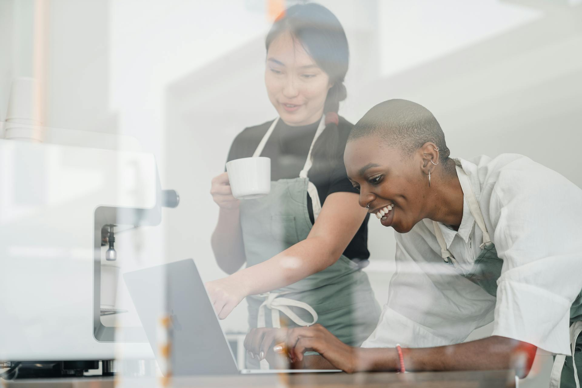 Through glass of friendly young Asian head barista showing young cheerful African American female coworker how to use special program on laptop