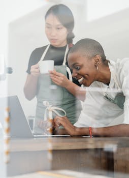 Two baristas having a fun coffee break while browsing a laptop indoors, enjoying a relaxed workplace vibe.