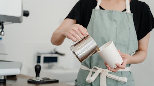 Crop unrecognizable female barista in apron pouring milk from steel jug into paper cup while working in modern coffeehouse