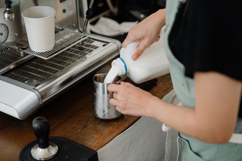 Free Side view of crop unrecognizable cafe worker pouring fresh milk into big stainless still cup while preparing beverage at counter with coffee machine in modern cafe Stock Photo
