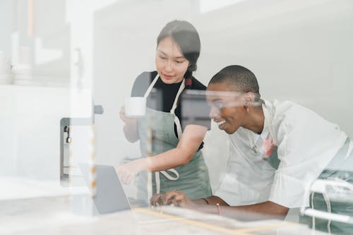 Young inexperienced African American female worker smiling and looking at laptop screen and Asian head barista explaining how special program works both shot through glass