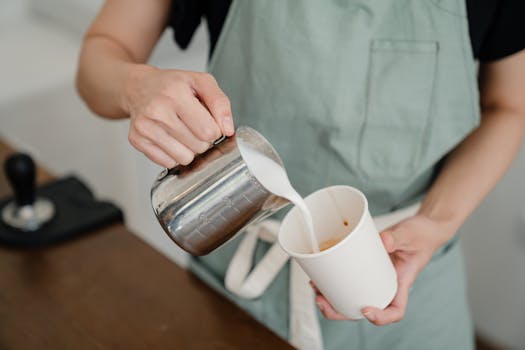 Barista preparing a hot drink by pouring milk into a coffee cup at a cafe.
