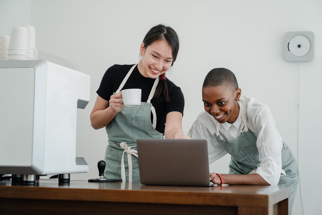 Happy Asian and black female colleagues working at coffee shop browsing net on laptop and smiling while having break at work