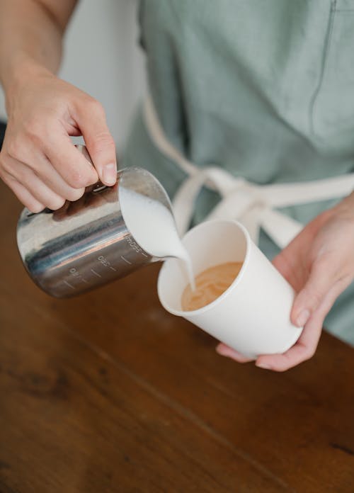 Crop faceless barista pouring whipped milk in paper coffee cup to make delicious latte