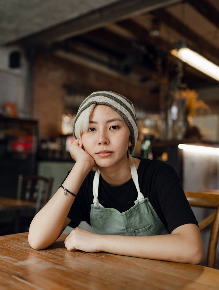 Female Barista In Beanie And Apron Resting Chin On Had