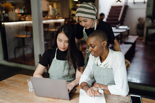 Free Smiling multiracial female coworkers in casual wear and aprons using modern netbook while working on project together in creative cafe Stock Photo
