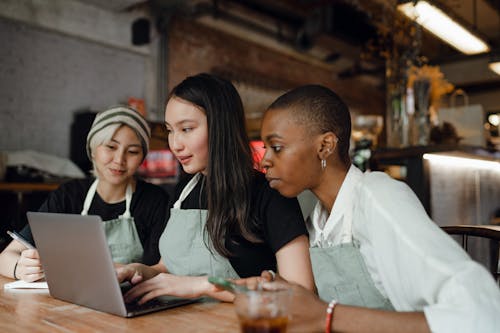 Content diverse women in aprons browsing laptop in cafeteria