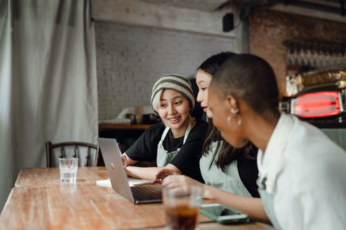 Free Happy smiling multiracial coworkers in aprons sitting at shabby table with drinks and working together on modern netbook in creative workspace Stock Photo