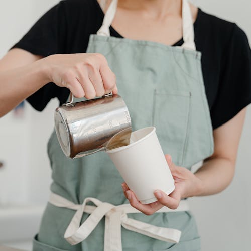 Crop barista in apron pouring milk into paper cup