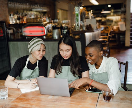 Happy smiling multiethnic waitresses in aprons watching tutorials on netbook while gathering at shabby table with notebook and drinks in cozy creative cafe