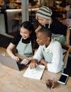 Astonished surprised young diverse waitresses in aprons gathering at wooden table with netbook and browsing internet while taking notes and working in creative workplace