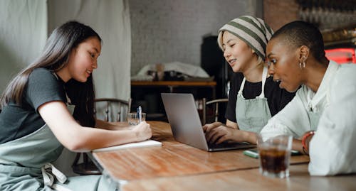 Free Positive smiling multiethnic female coworkers wearing casual clothes and aprons sitting at wooden table with modern laptop and taking notes while working on project together in creative workplace Stock Photo