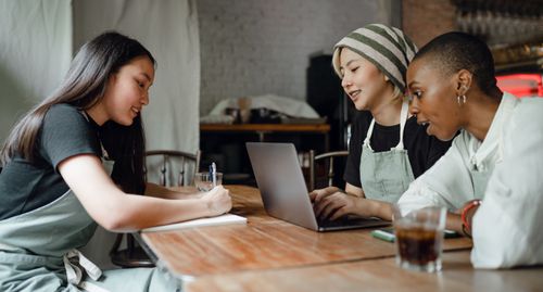 Free Positive smiling multiethnic female coworkers wearing casual clothes and aprons sitting at wooden table with modern laptop and taking notes while working on project together in creative workplace Stock Photo