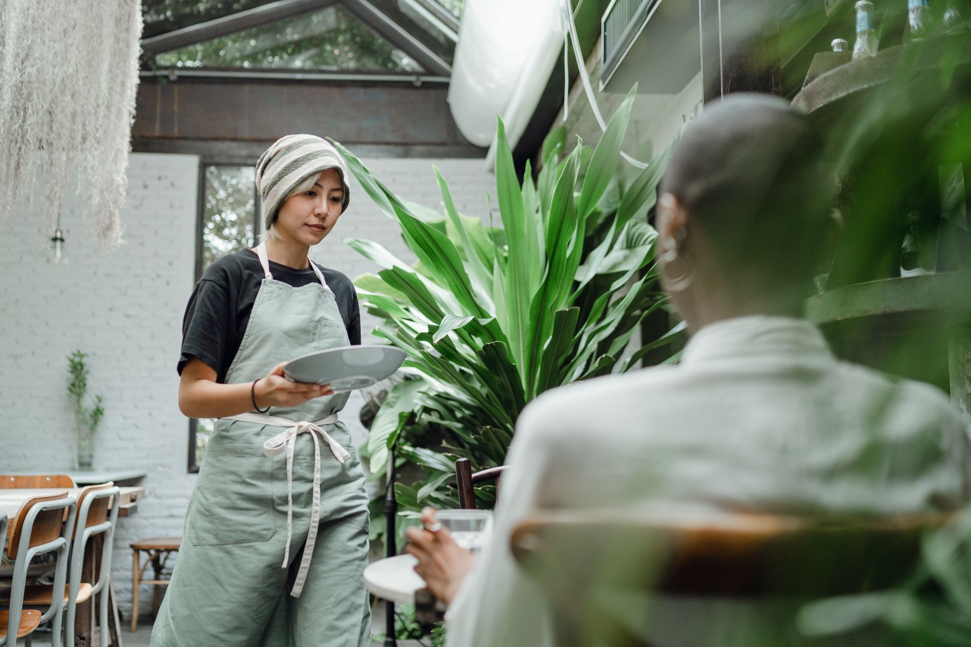 Positive young ethnic waitress in hat and apron carrying plate with meal and serving female guest while working in creative restaurant full of lush green plants