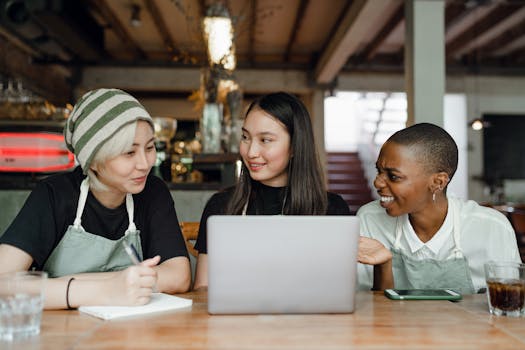 Smiling positive multiracial coworkers wearing aprons browsing modern netbook and having conversation while sitting at wooden table in cozy cafe