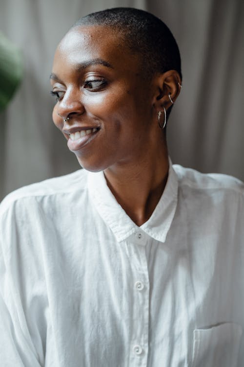 Portrait of happy smiling bald African American female with earrings wearing white casual blouse and looking away dreamily while standing against blurred background