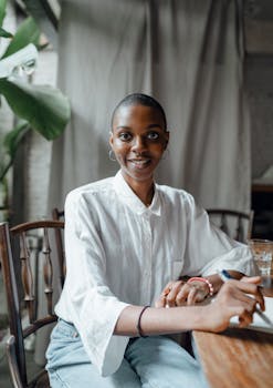 A cheerful woman with a bald hairstyle writing in a notebook at a wooden table indoors.