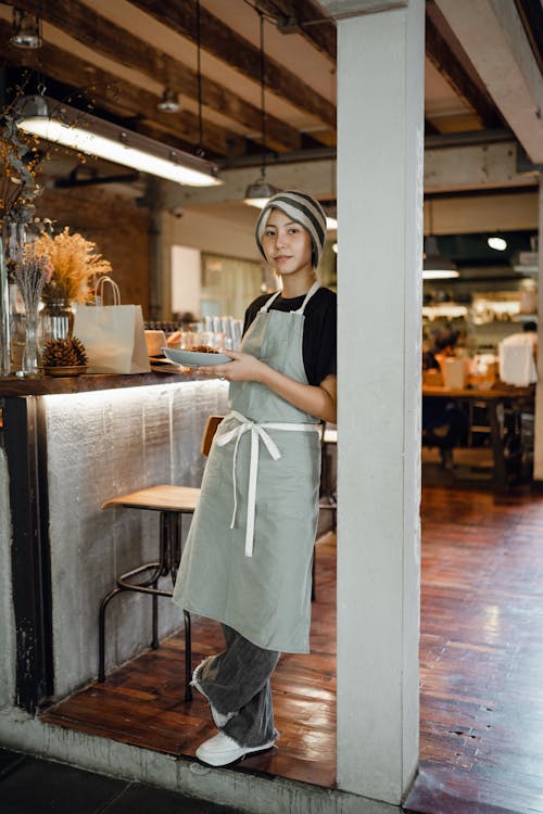 Positive young Asian waitress in apron and hat with plate leaning on beam and looking at camera before serving food in cozy modern restaurant