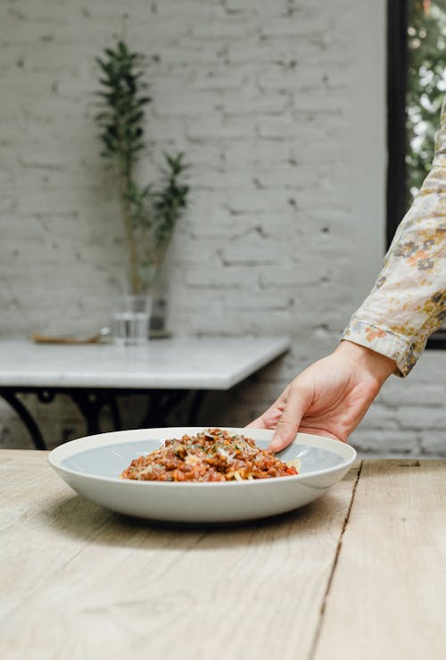 Crop unrecognizable female putting bowl with yummy fresh pasta with tomato sauce on wooden shabby table against decorative white brick wall in restaurant
