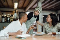 Joyful diverse restaurant critics tasting food in cozy cafe and taking notes in notepad while sitting at wooden table and talking to smiling waitress
