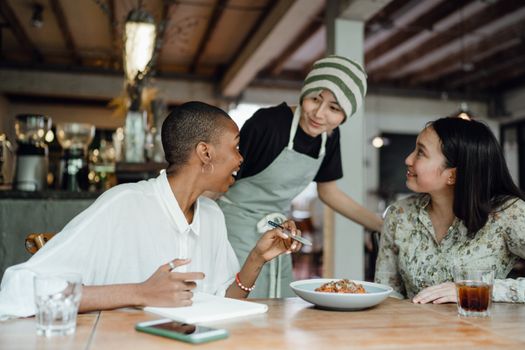 Joyful diverse restaurant critics tasting food in cozy cafe and taking notes in notepad while sitting at wooden table and talking to smiling waitress