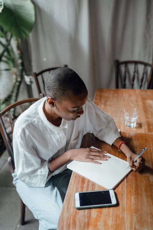 From above of black left handed female in casual wear taking notes in notepad while sitting at shabby table with smartphone against gray curtains