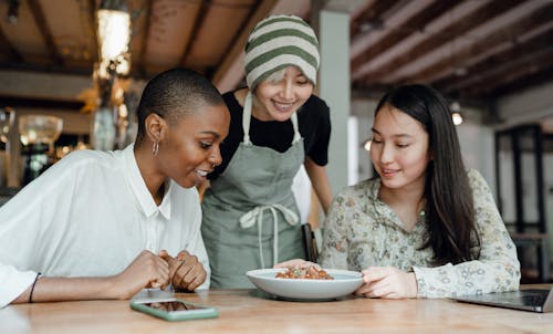 Free Smiling positive multiracial females in casual clothes sitting at table in modern cafe near cheerful waitress and looking at delicious dish Stock Photo