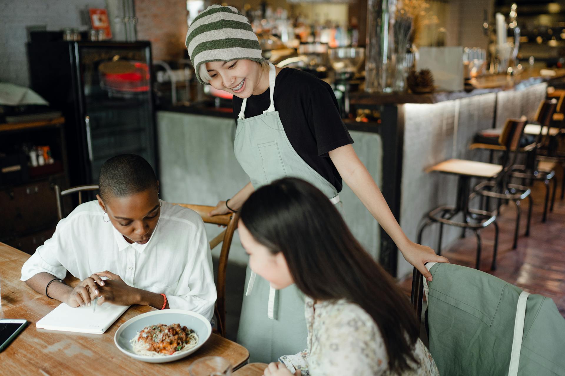 Positiv ung asiatisk servitris i förkläde och hatt som serverar läckra pasta för intresserade olika kvinnliga kunder i mysigt café