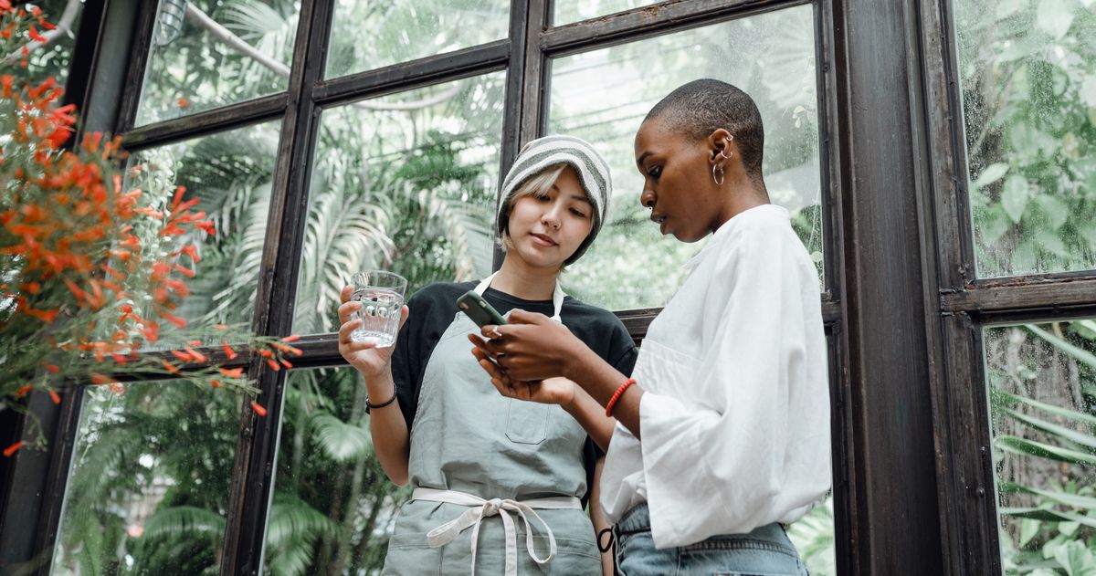 Two women standing by a greenhouse window looking at a smartphone together
