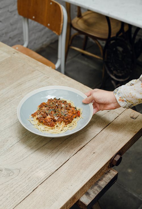 From above of crop anonymous young female serving plate of delicious pasta bolognese on wooden table in outdoor cafe