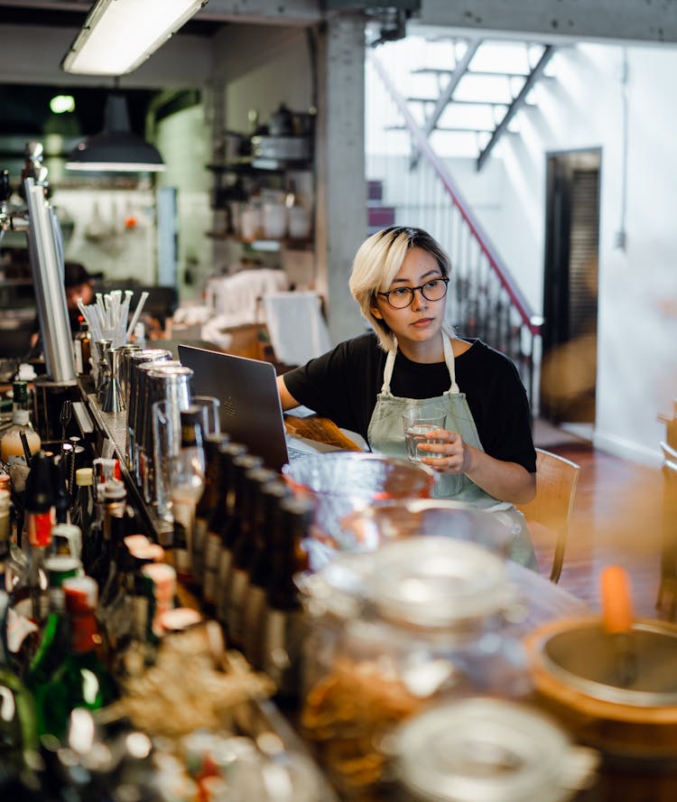 Focused Young Ethnic Female Bartender Drinking Water At Counter In Pub