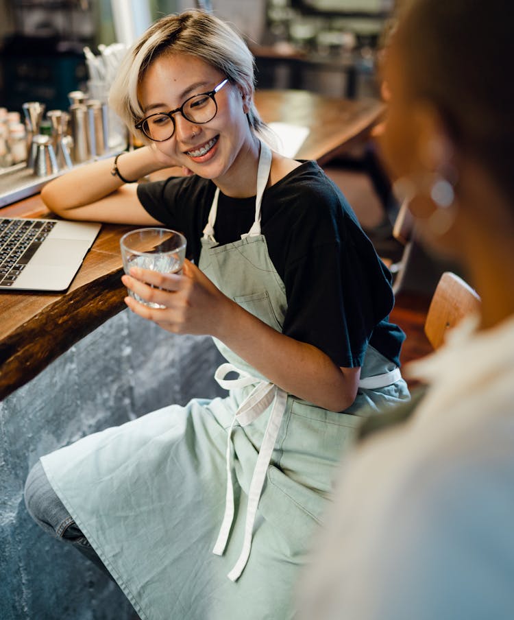 Cheerful Young Asian Woman Sitting Leaning On Bar Counter With Glass Of Water