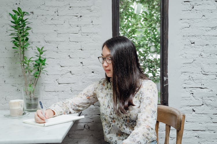 Young Female Asian Student Taking Notes In Planner Sitting At Table