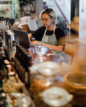 A focused young Asian female bartender uses her laptop while working in a bustling bar.
